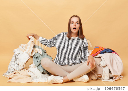 Shocked Caucasian woman posing near heap of multicolored unsorted clothes isolated over beige background sorting used clothing looking at camera with open mouth. Shocked Caucasian woman posing near heap of multicolored unsorted clothes isolated over beige background sorting used clothing looking at camera with open mouth. 106476547