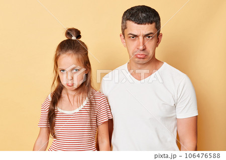 Unhappy sad upset man and little kid girl standing together isolated over beige background looking at camera with pout lips being offended expressing negative feelings 106476588