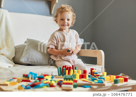 Wooden construction set. Montessori learning approach. Cognitive development puzzles. Happy smiling little blonde baby girl palying with wooden sorter toys at home interior. 106476771