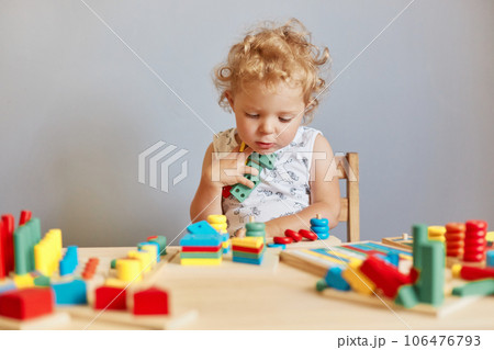 Creative learning moments. Early education. Nursery's fun-filled activities. Pensive wavy haired blonde toddler baby playing wooden educational toys isolated over gray background 106476793