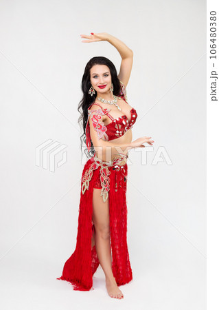 A brunette girl in a red dance costume performs oriental dances, a gray background A brunette girl in a red dance costume performs oriental dances, a gray background 106480380