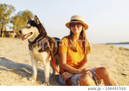 Woman in hat and sunglasses sits with her dog on the beach and enjoying sunset Woman in hat and sunglasses sits with her dog on the beach and enjoying sunset 106481588