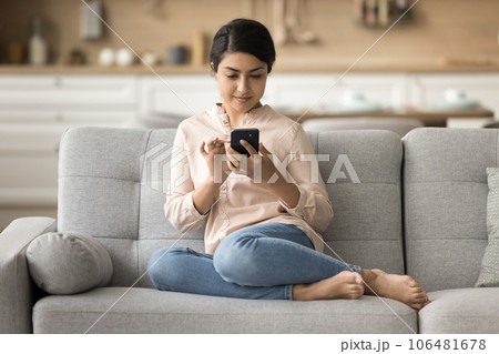 Positive pretty Indian woman resting on home sofa with smartphone Positive pretty Indian woman resting on home sofa with smartphone 106481678