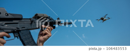 A man aims to shoot a rifle at a flying drone against a blue sky.  106483553