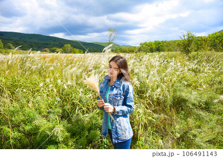Young smiling woman holding bouquet of Miscanthus sinensis (Chinese silver grass) in her hands and looking at it against the background of a field. Autumn in Russia. Copy space Young smiling woman holding bouquet of Miscanthus sinensis (Chinese silver grass) in her hands and looking at it against the background of a field. Autumn in Russia. Copy space 106491143