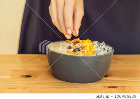 Woman cooks smoothie bowls made with mango, banana, granola, grated coconut, dragon fruit, chia seeds and mint on wooden background. Concept fruits, vitamins 106496839
