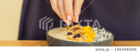 Woman cooks smoothie bowls made with mango, banana, granola, grated coconut, dragon fruit, chia seeds and mint on wooden background. Concept fruits, vitamins BANNER, LONG FORMAT 106496840