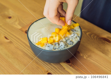 Woman cooks smoothie bowls made with mango, banana, granola, grated coconut, dragon fruit, chia seeds and mint on wooden background. Concept fruits, vitamins Woman cooks smoothie bowls made with mango, banana, granola, grated coconut, dragon fruit, chia seeds and mint on wooden background. Concept fruits, vitamins 106496841