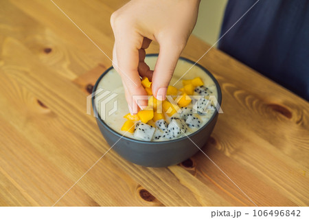 Woman cooks smoothie bowls made with mango, banana, granola, grated coconut, dragon fruit, chia seeds and mint on wooden background. Concept fruits, vitamins 106496842