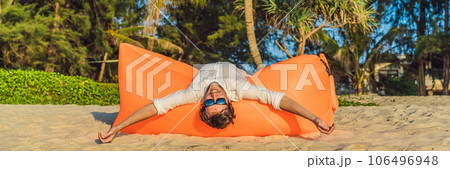 Summer lifestyle portrait of man sitting on the orange inflatable sofa on the beach of tropical island. Relaxing and enjoying life on air bed BANNER, LONG FORMAT 106496948