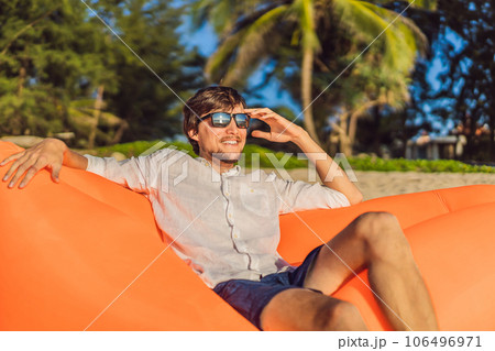 Summer lifestyle portrait of man sitting on the orange inflatable sofa on the beach of tropical island. Relaxing and enjoying life on air bed Summer lifestyle portrait of man sitting on the orange inflatable sofa on the beach of tropical island. Relaxing and enjoying life on air bed 106496971