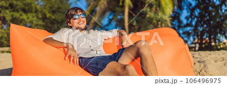 Summer lifestyle portrait of man sitting on the orange inflatable sofa on the beach of tropical island. Relaxing and enjoying life on air bed BANNER, LONG FORMAT Summer lifestyle portrait of man sitting on the orange inflatable sofa on the beach of tropical island. Relaxing and enjoying life on air bed BANNER, LONG FORMAT 106496975