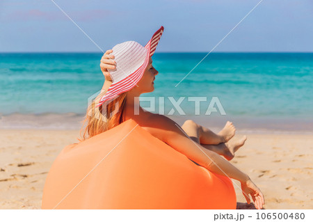 Summer lifestyle portrait of pretty girl sitting on the orange inflatable sofa on the beach of tropical island. Relaxing and enjoying life on air bed 106500480