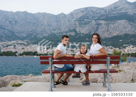 Portrait of happy family sits on bench on sea coast and look at the camera. Back view of sea and mountains Portrait of happy family sits on bench on sea coast and look at the camera. Back view of sea and mountains 106503330