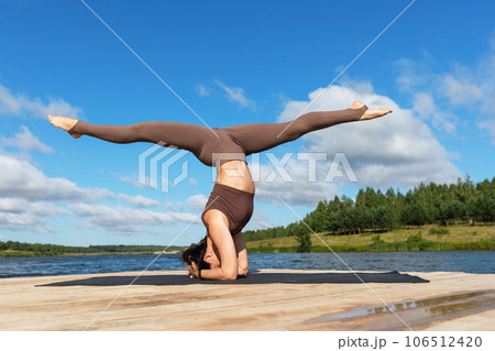A woman practicing yoga performs the Sirshasana exercise with Hanumanasana, an inverted asana with a longitudinal twine, trains in sportswear on the shore of a lake on a warm sunny morning 106512420