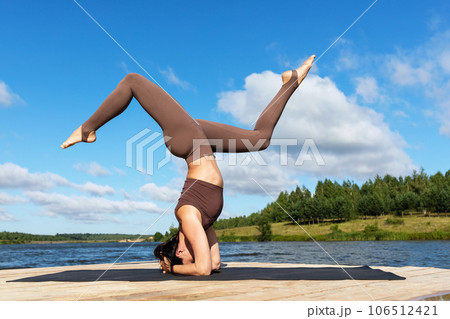 A woman practicing yoga, performs a variation of the Shirshasana exercise, inverted asana, headstand and handstand, trains in sportswear on the shore of a lake on a warm sunny morning 106512421