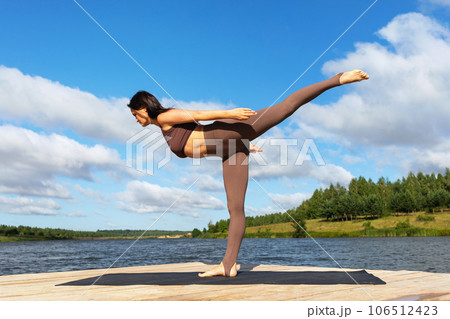 A woman practicing yoga, doing a variation of the Virabhadrasana exercise, the number three warrior pose, is training in sportswear on the shore of a lake on a warm sunny morning 106512423