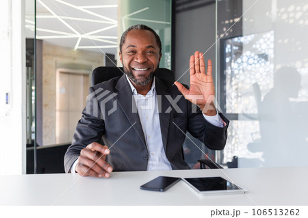 Webcam view, video call online meeting, man looking at camera waving hand talking to colleagues and greeting, businessman at workplace, on remote meeting in office interior in business suit boss. Webcam view, video call online meeting, man looking at camera waving hand talking to colleagues and greeting, businessman at workplace, on remote meeting in office interior in business suit boss. 106513262