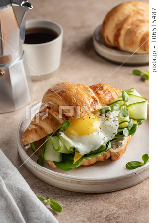 Croissant sandwich with fried egg, cheese, cucumber and micro greens and coffee over beige background. Healthy delicious breakfast Croissant sandwich with fried egg, cheese, cucumber and micro greens and coffee over beige background. Healthy delicious breakfast 106515487