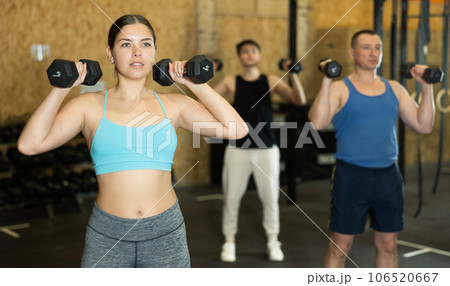 Young dedicated active strong fit woman doing exercises for shoulders while pushing up dumbbells in CrossFit center Young dedicated active strong fit woman doing exercises for shoulders while pushing up dumbbells in CrossFit center 106520667