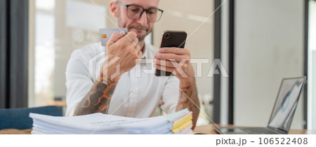 Stressed caucasian male business holding credit card making online payment using smartphone at office Stressed caucasian male business holding credit card making online payment using smartphone at office 106522408