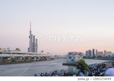 【日本】夕方の汐入公園の土手で隅田川花火大会の開始を待つ群衆とスカイツリーのある風景 106525649