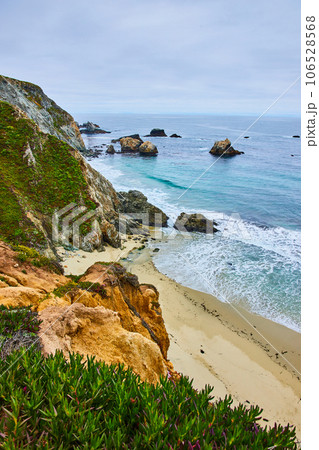 Scenic green cliffy hillside with sandy shore below and ocean waves crashing into shoreline 106528568