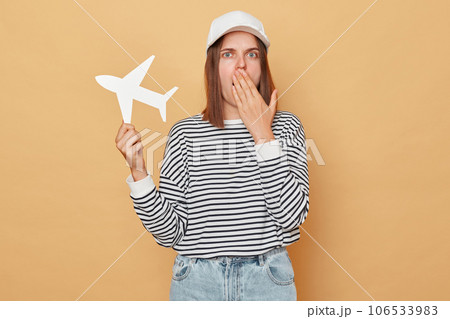 Shocked woman wearing baseball cap and striped shirt holding white paper plane isolated over beige background booking wrong direction covering mouth with palm Shocked woman wearing baseball cap and striped shirt holding white paper plane isolated over beige background booking wrong direction covering mouth with palm 106533983