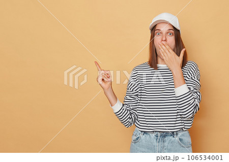 Surprised amazed woman wearing baseball cap and striped shirt isolated over beige background covering mouth with palm pointing at advertisement area space for content 106534001