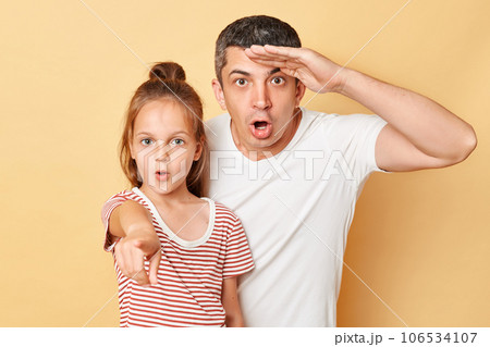 Shocked father with hand near forehead and daughter pointing at camera family wearing casual t-shirts standing isolated over beige background. 106534107