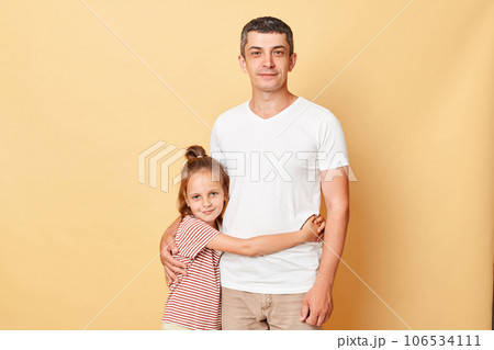 Portrait of family father and daughter wearing casual t-shirts standing isolated over beige background kid and dad hugging each other looking at camera. Portrait of family father and daughter wearing casual t-shirts standing isolated over beige background kid and dad hugging each other looking at camera. 106534111