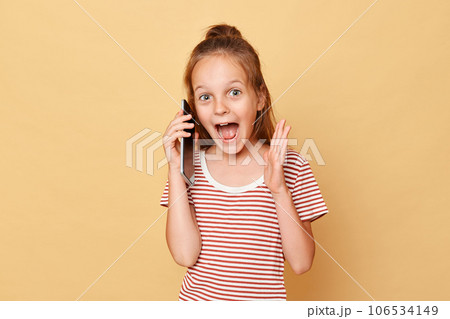 Amazed excited little brown haired girl wearing striped t-shirt standing isolated over beige background talking on mobile phone screaming hearing excellent news. Amazed excited little brown haired girl wearing striped t-shirt standing isolated over beige background talking on mobile phone screaming hearing excellent news. 106534149
