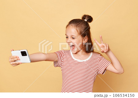Overjoyed little brown haired girl wearing striped t-shirt standing isolated over beige background making selfie on smart phone showing v sign broadcasting livestream in kids vlog. Overjoyed little brown haired girl wearing striped t-shirt standing isolated over beige background making selfie on smart phone showing v sign broadcasting livestream in kids vlog. 106534150