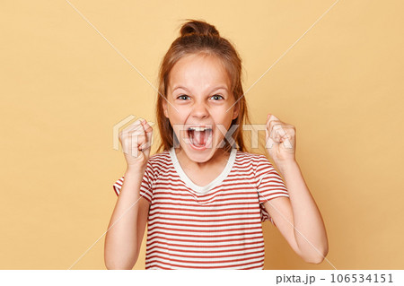 Extremely happy crazy little brown haired girl wearing striped t-shirt standing isolated over beige background clenched fists with excitement celebrating holiday start. Extremely happy crazy little brown haired girl wearing striped t-shirt standing isolated over beige background clenched fists with excitement celebrating holiday start. 106534151