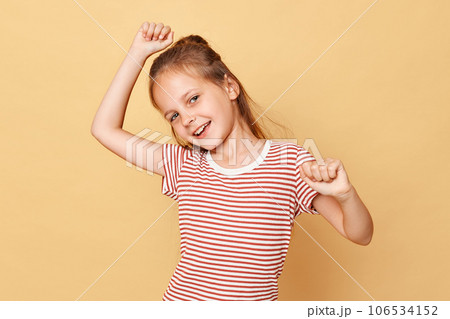 Cheerful happy little brown haired girl wearing striped t-shirt standing isolated over beige background dancing with raised arms looks overjoyed and expressing happiness. Cheerful happy little brown haired girl wearing striped t-shirt standing isolated over beige background dancing with raised arms looks overjoyed and expressing happiness. 106534152