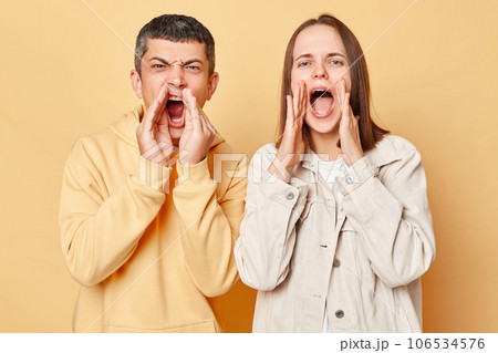 Excited woman and man wearing casual style clothing standing isolated over beige background screaming loud with hands near mouths saying important information. 106534576