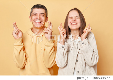 Hopeful woman and man wearing casual style clothing standing isolated over beige background making wishtogetherposing with crossed fingers dreaming for good luck. 106534577