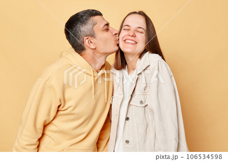 Loving couple woman and man wearing casual style clothing standing isolated over beige background expressing love, husband kissing his wife. Loving couple woman and man wearing casual style clothing standing isolated over beige background expressing love, husband kissing his wife. 106534598