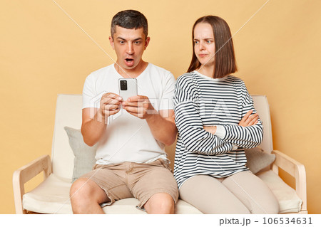 Shocked man using mobile phone and offended woman wearing casual clothing sitting on clothes isolated over beige background female feeling sadness and being lonely while husband involved in internet. Shocked man using mobile phone and offended woman wearing casual clothing sitting on clothes isolated over beige background female feeling sadness and being lonely while husband involved in internet. 106534631
