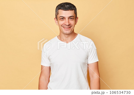 Smiling handsome man wearing white casual t-shirt standing isolated over beige background looking at camera being in good mood expressing positive emotions. Smiling handsome man wearing white casual t-shirt standing isolated over beige background looking at camera being in good mood expressing positive emotions. 106534730