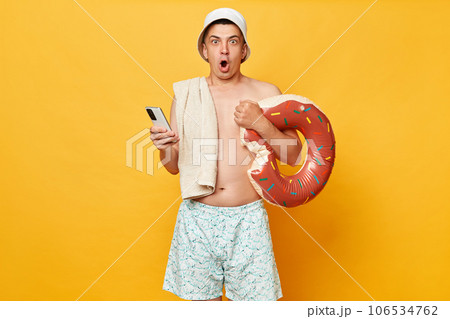 Shocked man wearing shorts swimsuit and panama holding donut rubber ring isolated on yellow background booking tickets back resting at tropical resort. 106534762