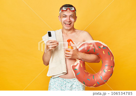 Smiling positivejoyful tourist man at summer resort standing with rubber ring isolated over yellow background using mobile phone while relaxing near the sea. Smiling positivejoyful tourist man at summer resort standing with rubber ring isolated over yellow background using mobile phone while relaxing near the sea. 106534785