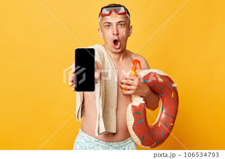Shocked surprised tourist man at summer resort standing with rubber ring isolated over yellow background showing cell phone with empty screen copy space for advertisement. Shocked surprised tourist man at summer resort standing with rubber ring isolated over yellow background showing cell phone with empty screen copy space for advertisement. 106534793