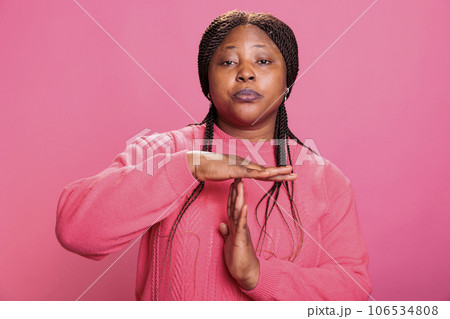 Angry woman doing timeout gesture with t shape hands making break time sign with arms in studio with pink background. African american young adult advertising pause symbol with palms Angry woman doing timeout gesture with t shape hands making break time sign with arms in studio with pink background. African american young adult advertising pause symbol with palms 106534808