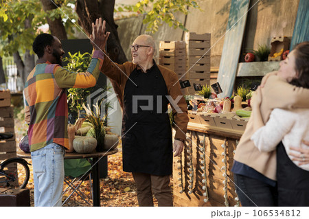 Cheerful senior man giving high five to regular customer while selling fresh organic vegetables and fruits at farmers market, local vendors greeting consumers with friendly smile at greenmarket 106534812