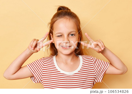 Dreamy cheerful dark haired little girl showing v-sign near eyes winking wearing casual style striped T-shirt standing isolated over beige background. 106534826