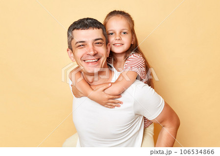 Smiling joyful family father and daughter wearing casual t-shirts standing isolated over beige background looking at camera with positive expressionpiggyback. Smiling joyful family father and daughter wearing casual t-shirts standing isolated over beige background looking at camera with positive expressionpiggyback. 106534866