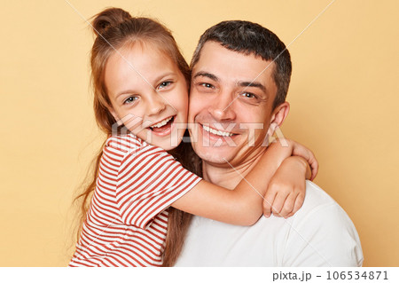 Joyful family father and daughter wearing casual t-shirts standing hugging each other looking at camera with happy faces isolated over beige background 106534871