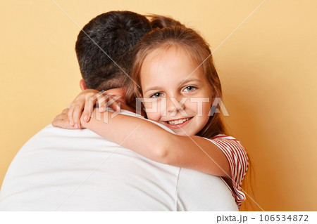 Father posing backwards embracing his daughter wearing casual t-shirts standing isolated over beige background kid looking smilign ar camera parent love. 106534872