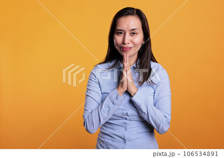 Positive asian woman praying to god in front of camera, holding hands in prayer gesture and asking for good luck. Religious model expressing hope and belief, standing in studio over yellow background Positive asian woman praying to god in front of camera, holding hands in prayer gesture and asking for good luck. Religious model expressing hope and belief, standing in studio over yellow background 106534891